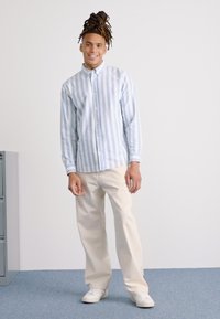 Camisa abotonada a rayas azul claro y blanca con mangas largas, combinada con pantalones beige y zapatillas blancas, sobre un fondo neutro.