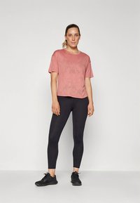 Rosa treningst-skjorte med avslappet passform, kombinert med svarte tights. Stoffet virker mykt med en svak glans. Svarte treningssko.