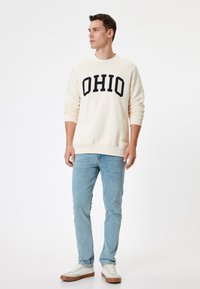 Krämfärgad sweatshirt med "OHIO" i fet svart text, tillsammans med ljusblå jeans och vita sneakers med bruna accenter. Slät textur.