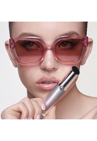 Roze oversized zonnebril met gradient lenzen, glanzende nude lippen, houdt een zilveren cosmetische tube vast met een zwarte applicatortip.