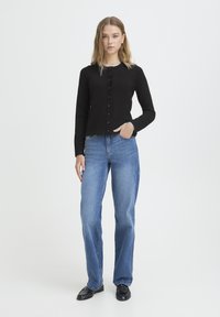 Črni dolg rokav, gumb na srajčni modrček v kombinaciji s svetlo modrimi kavbojkami s širokimi hlačnicami in črnimi loaferji, postavljeno na belem ozadju.