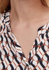 Bunt gemusterte Bluse mit geometrischem Muster in Marineblau, Weiß und Rost. V-Ausschnitt-Design mit Knöpfen und strukturiertem Stoff.