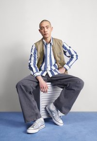 Jeune homme assis sur une valise argentée, portant une chemise rayée bleu et blanc, un gilet beige, un pantalon large gris et des baskets grises, sur un sol bleu.