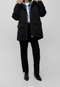 Parka nera con cappuccio foderato di pelliccia, dotata di zip frontale, due tasche inferiori e una vestibilità leggermente oversize, abbinata a pantaloni neri e stivali con tacco.