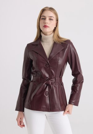 Femme portant une veste en cuir bordeaux ceinturée par-dessus un pull à col roulé beige et un pantalon blanc, debout devant un fond blanc uni.
