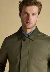 Vīrietis ar cirtainiem matiem, ģērbies olīvu zaļā žaketē virs zila kreklā ar apkakli, stāv uz priekšu pret bēšainu fona.