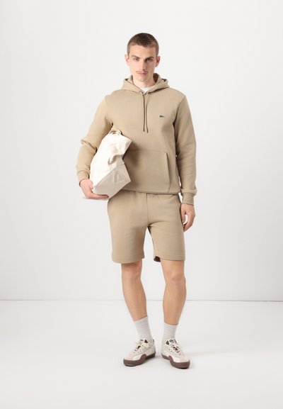 Sweatshirt à capuche en coton beige avec poche kangourou, associé à un short assorti. Le modèle tient un sac crème. Chaussettes blanches texturées et baskets.