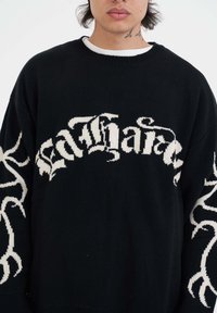 Czarny sweter dzianinowy z przeszłymi białymi literami oraz dekoracyjnymi białymi wzorami na rękawach. Okrągły dekolt, luźny krój.