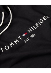Sudadera negra con texto bordado en blanco que dice "TOMMY HILFIGER" y un pequeño logo tricolor. Incluye capucha con cordón. Textura de tela suave.