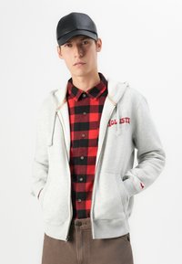 Giovane uomo con un berretto nero, una camicia a scacchi rossa e nera, una felpa grigia chiara con cerniera e pantaloni marroni, in piedi con le mani nelle tasche.