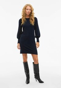 Abito in maglia blu navy con maniche lunghe a palloncino, scollo arrotondato e vestibilità rilassata. Indossato con stivali neri alti con punta appuntita.