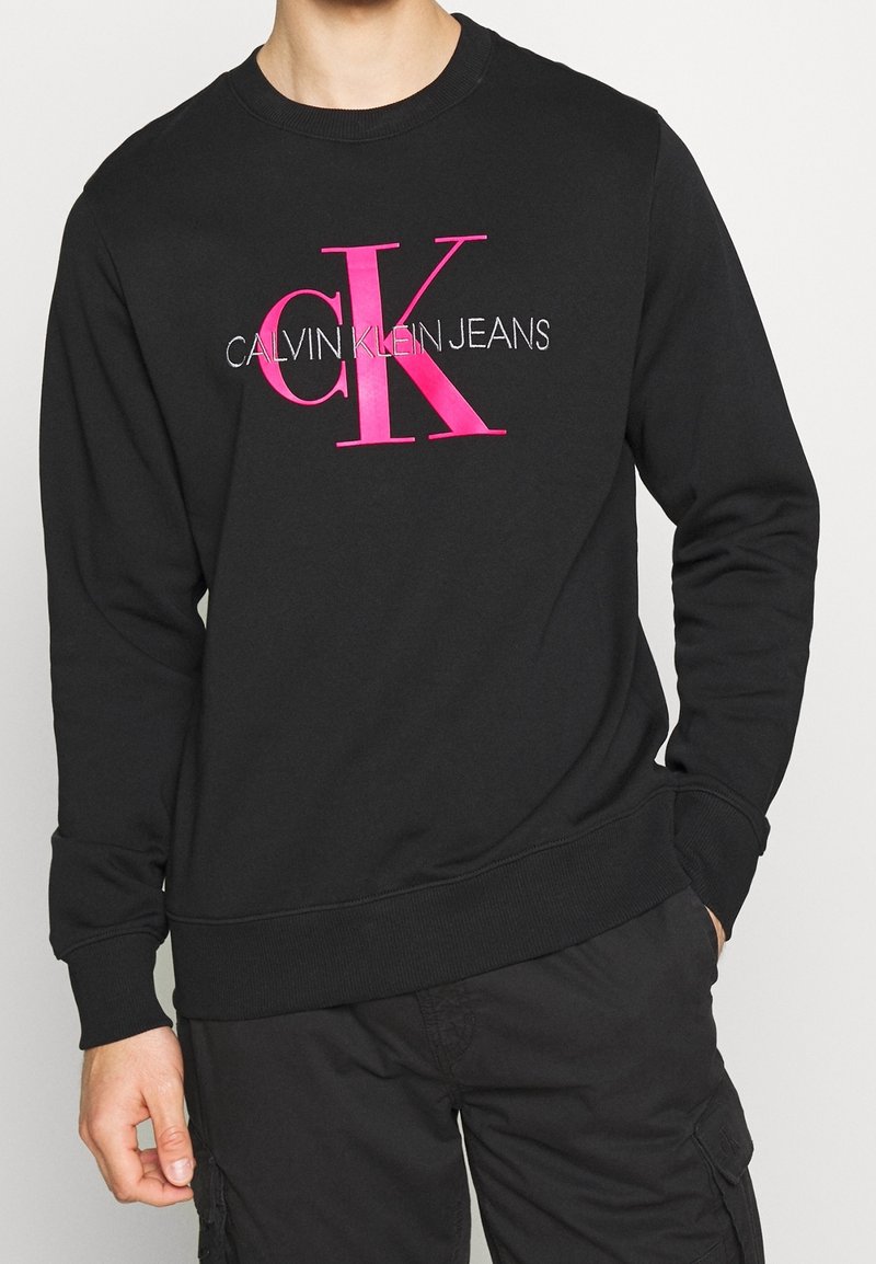 Sweatshirt noir à col rond en coton, avec un grand logo "CK" rose et le texte "CALVIN KLEIN JEANS" brodé en blanc.