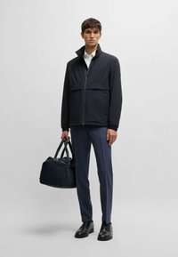 Blå jacka med dragkedja framtill och hög krage, i kombination med skräddade byxor och en mörk väska. Slät tyg och strukturerad design.