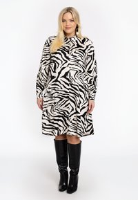 Zebra-Print Hemdkleid in Creme und Schwarz, mit langen Ärmeln, Knopfschluss und lockerer Passform. Kombiniert mit kniehohen schwarzen Stiefeln.