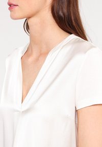 Donna che indossa una blusa bianca in satin a maniche corte con scollatura a V, mostrata dalla zona del collo fino alla parte centrale del torso, con lunghi capelli castani visibili.