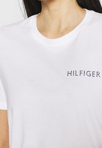 Bílé bavlněné tričko s kulatým výstřihem a modrým hroty zdobeným logem "HILFIGER" na levé části hrudi. Hladká textura, klasický střih.