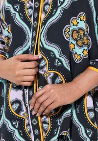 Schwarze Bluse aus Stoff mit kunstvollen blauen, orangefarbenen und lila Blumenmustern, mit Knopfdetails und kontrastierendem Saum.