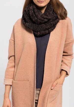 Femme portant un manteau beige aux manches retroussées et grandes poches frontales, un haut noir en dessous, et une grosse écharpe tricotée brun foncé.