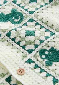 Tissu crocheté avec des motifs géométriques et floraux verts et blancs, orné d'un bouton marron à quatre trous sur un fil texturé.