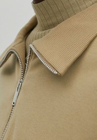 Beżowa kurtka na zamek błyskawiczny z prążkowanym wysokim kołnierzem, srebrnymi detalami zamka i gładką teksturą tkaniny. Zbliżenie ukazujące cechy designu.