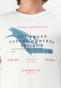 Hvit bomulls t-skjorte med blå og rød tekst med luftfartstemaer, inkludert grafikk av et fly og tekniske termer.