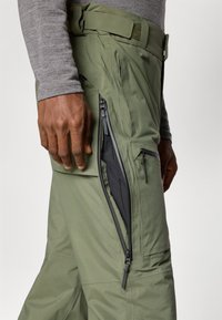 Chaki žalio atspalvio lauko kelnių su užtrauktuku kišenėje ant šlaunies. Ypatybės: tvirta medžiaga, reguliuojamas diržas ir elegantiškas dizainas.