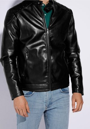 Mann trägt schwarze Lederjacke über grünem Hemd und hellblauer Jeans, steht mit entspannten Händen an den Seiten.