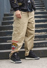 Beige cargobyxor med två sidofickor, prydda med en broderad tigerdesign och röd text. Matchas med svarta hög-top sneakers.