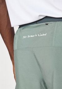 Pantalones deportivos de color verde claro con una tela texturizada y una inscripción bordada "Ningún humano es limitado" en la parte trasera del cinturón.