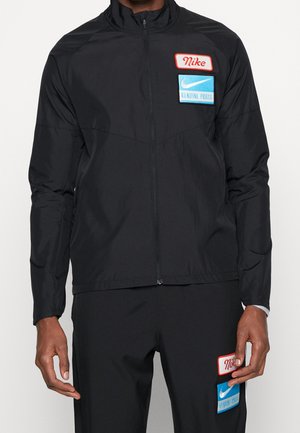 Homme portant un survêtement noir Nike avec une veste zippée et un pantalon assorti, tous deux ornés de patchs Nike rouges et bleus.