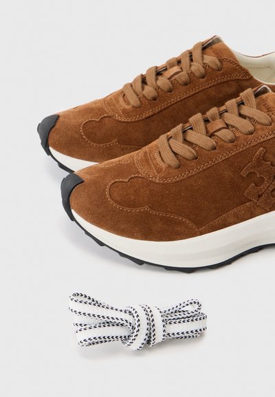 Sneakers din suede maro cu sireturi maro cu model și o pereche extra de sireturi spirale albe și negre pe un fond alb.