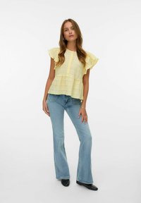 Geel rimpelblouse met een textuurontwerp en kleine knopen, gecombineerd met lichtblauwe flared jeans. Zwart schoeisel maakt de outfit compleet.