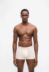 Μυώδης άνδρας όρθιος, φοράει ανοιχτό μπεζ ανδρικά εσώρουχα τύπου μποξέρ, μπροστά σε απλό λευκό φόντο, κοιτάζει μπροστά με ουδέτερη έκφραση.