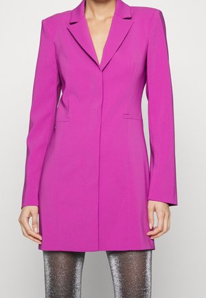 Woman wearing a bright pink long blazer over sparkly silver leggings, standing against a plain light background.