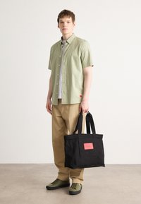 Bolso de lona negro con una etiqueta roja, llevado por una persona con una camisa de manga corta verde claro y pantalones beiges, que lleva zapatillas verdes.