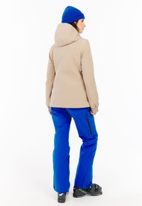 Beige waterdichte jack met capuchon, gedragen over blauwe broek. Heeft elastische manchetten en zijzakken, met zwarte ritsen en zwarte skikaplaarzen.