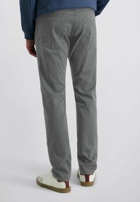 Uomo che indossa pantaloni grigi a gamba dritta, sneakers bianche con dettaglio del tallone marrone, e un maglione blu, in piedi contro uno sfondo bianco semplice.