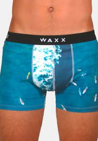 Shorts de bain bleus avec un motif de vague océanique et de planche de surf. Taille élastique noire avec le logo "WAXX" ; texture lisse et coupe ajustée.
