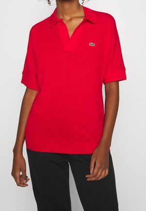 Mujer vistiendo una camiseta tipo polo de manga corta de color rojo brillante con un pequeño logo de cocodrilo en el pecho y pantalones negros, sobre un fondo liso.