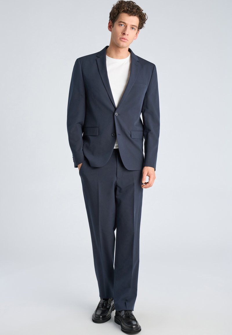 Ung mann står iført en mørk blå dress med hvit skjorte og svarte selskapssko foran en ensfarget lys bakgrunn.