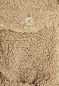 Tissu en peluche beige avec une surface texturée et bouclée. Présente un seul bouton crème permettant de fixer un rabat. Matériau doux et épais.