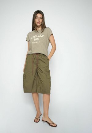 Kvinde iført beige, stribet "Atelier 2010 Mos Mosh" t-shirt, olivengrøn knælange cargobukser og brune sandaler med lav hæl, stående op ad en hvid væg.