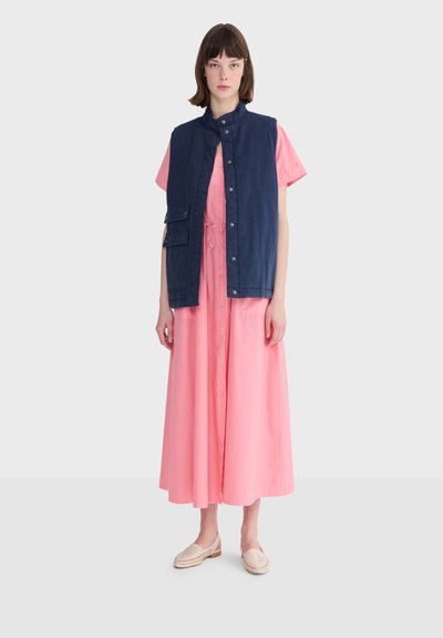 Femme portant une longue robe rose boutonnée, un gilet sans manches bleu marine avec des poches, et des mocassins tissés beiges, debout devant un fond uni.