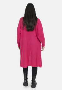 Lång, öppet stickad kofta i knallrosa, med vida ärmar och avslappnad passform, ihop med svarta leggings och kraftiga svarta stövlar.