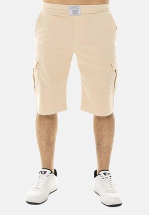 Beige cargoshorts met zijzakken gedragen door een staande persoon gecombineerd met witte en zwarte sneakers op een witte achtergrond.