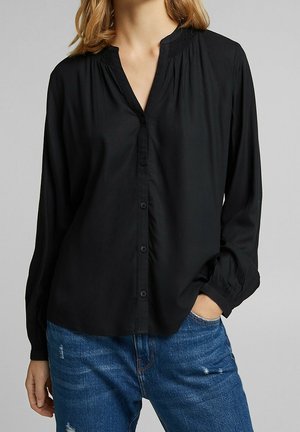Sort langærmet skjorte med knaplukning foran, v-hals og sammensyning ved halsudskæringen. Båret med blå slidte jeans.