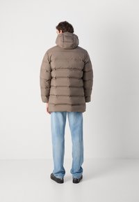 Een taupe pufferjack met een verticale quilted ontwerp, een capuchon en elastische manchetten, gecombineerd met lichtblauwe jeans en zwarte schoenen.