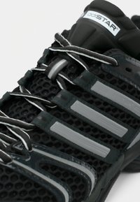 Chaussure de sport noire avec une tige en mesh, des bandes grises réfléchissantes et une semelle texturée. Fermeture à lacets avec des accents gris et blancs.