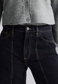 Fekete farmernadrág látható varrással, gombos záródással és két elülső zsebbel, szürke, texturált kötött anyagból készült kardigánnal párosítva.