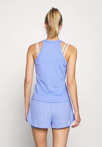 Donna con coda di cavallo bionda, indossa una canotta atletica senza maniche di colore azzurro chiaro e pantaloni corti coordinati, in piedi di fronte a uno sfondo bianco semplice.